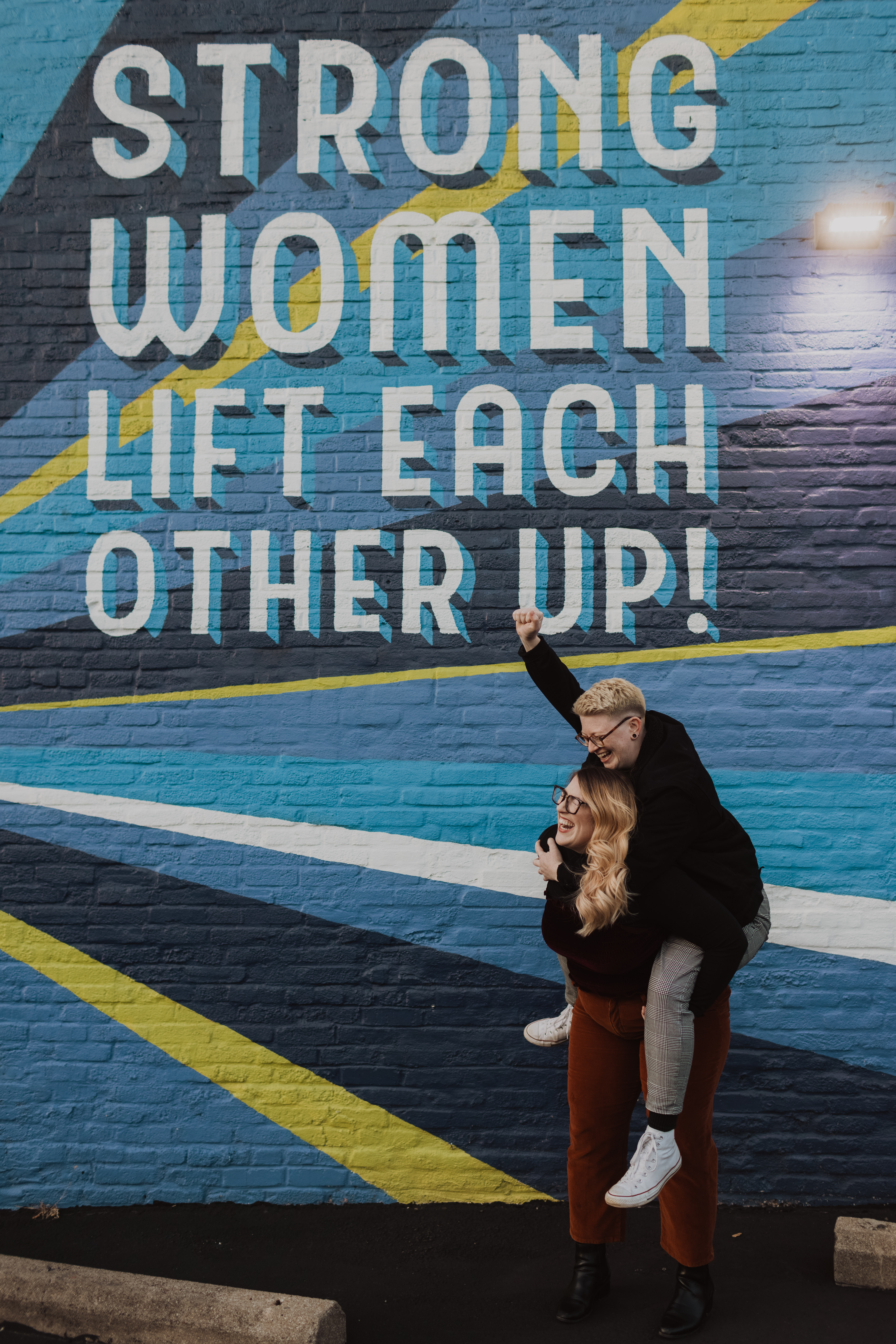 Tori Lumen is on Chelsie Lumen's back with her fist raised in front of a Strong Women Life Each Other Up mural